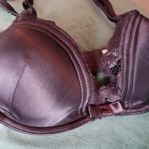 May include: A dark purple satin bra with lace detailing. The bra has adjustable straps and a hook and eye closure.