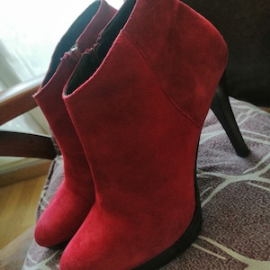Peut inclure: Une paire de bottines en daim rouge à talons noirs. Les bottines ont une pointe pointue et une fermeture éclair à l'intérieur de la cheville.