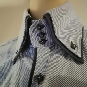 Puede incluir: Primer plano de una camisa a rayas con cuello doble. La camisa presenta un patrón de rayas azul claro y blanco, con ribetes y botones negros. El cuello es de color blanco contrastante.