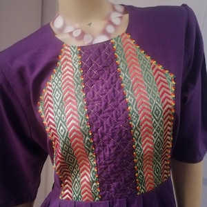 Könnte beinhalten: Lilafarbenes Kleid mit einem geometrischen Muster, das auf der Vorderseite gestickt ist. Die Stickerei ist in Rot-, Grün-, Gold- und Weißtönen gehalten.