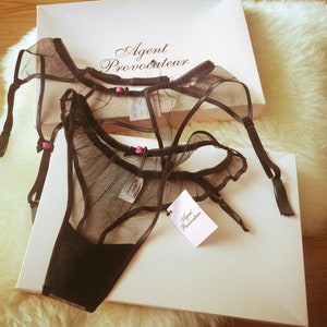 May include: Black lingerie set with delicate lace and pink accents. The set includes a suspender belt and a matching panty. The packaging features the Agent Provocateur logo.