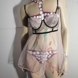 May include: A lingerie set on a mannequin. The set includes a light pink bra with black lace detailing, a sheer skirt, and polka dot underwear. The mannequin has body art and is wearing a silver bracelet.