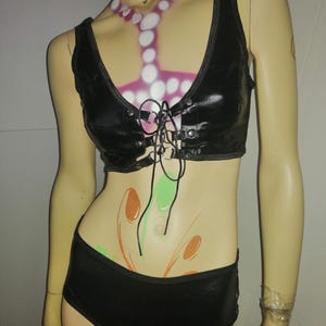 Peut inclure: Un ensemble noir deux pièces sur un mannequin. Le haut est un col en V avec un laçage sur le devant et des œillets argentés. Le short assorti est taille basse. Le mannequin a du body art rose, blanc, orange et vert.