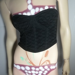 May include: A black strapless corset with a geometric pattern. The corset is on a mannequin with white polka dots painted on the torso.