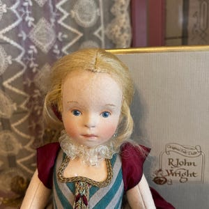 May include: A handcrafted doll with blonde hair and blue eyes, dressed in a teal, grey, and burgundy gown with a frilled collar. The doll has a detailed face and is a collectible item.