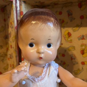 Puede incluir: Muñeca vintage con rostro pintado, ojos azules y vestido blanco con ribete de encaje. La muñeca tiene una línea de cabello marrón y posa con los brazos extendidos. El fondo incluye el interior estampado de un estuche vintage.