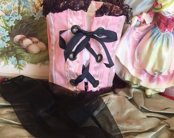 May include: A pink satin corset with black lace trim and black ribbon lacing. The corset has a bow at the back and is in a vintage style.