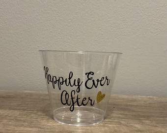 May include: A clear plastic cup with the words "Happily Ever After" in black script. A small gold heart is to the right of the text. The cup is sitting on a wooden surface.