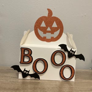 May include: A white Halloween gift box decorated with orange glitter. The box has the word "BOO" in orange letters with black outlines, and black bat cutouts. A glittery orange pumpkin is on top.