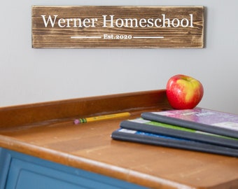 Puede incluir: Un letrero rectangular de madera marrón oscuro con texto blanco que dice Werner Homeschool Est.2020. El letrero está montado sobre un pupitre escolar de madera con base azul, con una manzana roja, un lápiz amarillo y cuadernos.