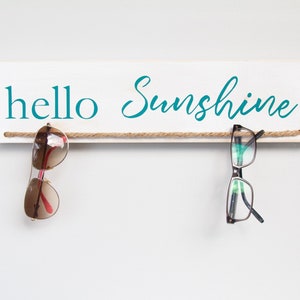 May include: White wooden sign with the text 'hello sunshine' in teal lettering. Two pairs of glasses hang from a rope attached to the sign. One pair is brown sunglasses and the other is black framed glasses.