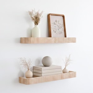 May include: Two light-colored wooden floating shelves with a vertical ribbed texture mounted on a white wall. The top shelf holds a cream-colored vase with dried flowers and a wooden picture frame. The bottom shelf holds two small vases with dried flowers and a stack of three books topped with a round stone sphere.