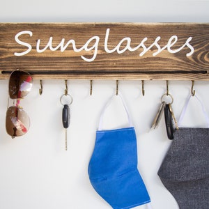 May include: A wooden wall-mounted organizer with the word "Sunglasses" written on it. It has six hooks for hanging items, including sunglasses, keys, and face masks. A pair of sunglasses, a set of keys, a blue face mask, and a gray face mask are hanging on the hooks.