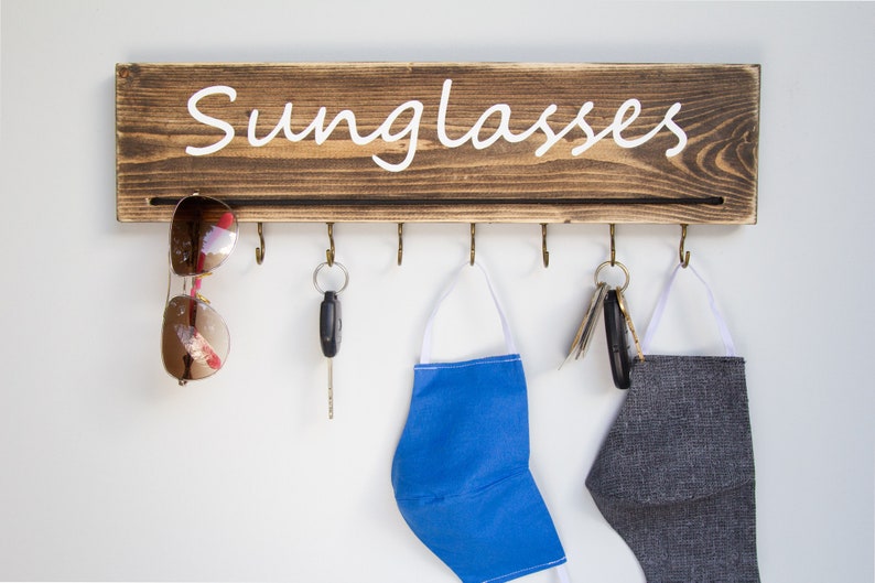 May include: A wooden wall-mounted organizer with the word "Sunglasses" written on it. It has six hooks for hanging items, including sunglasses, keys, and face masks. A pair of sunglasses, a set of keys, a blue face mask, and a gray face mask are hanging on the hooks.