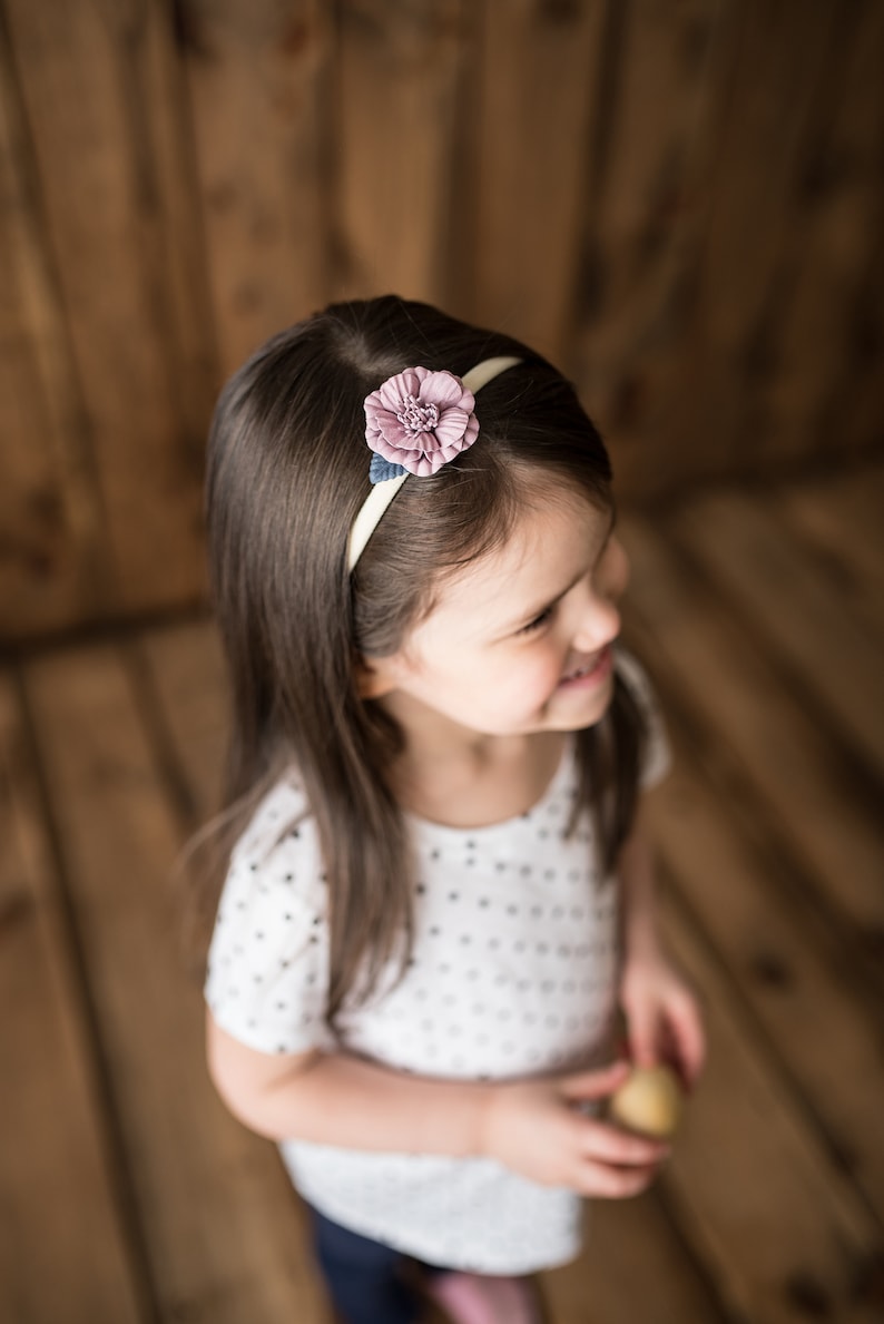 Pu&ograve; includere: Una bambina con lunghi capelli castani che indossa una camicia bianca a pois e una fascia per capelli con un fiore rosa.