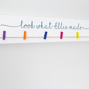 Op de afbeelding: Een wit houten bord met de tekst "look what Ellie made" in turquoise schrift. Een touwtje van touw is gespannen over het bord met gekleurde wasknijpers eraan bevestigd.