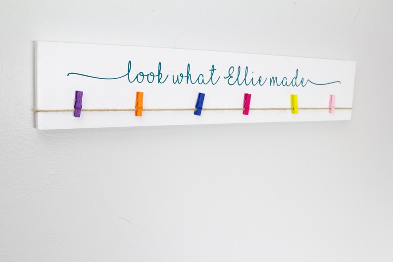 Op de afbeelding: Een wit houten bord met de tekst "look what Ellie made" in turquoise schrift. Een touwtje van touw is gespannen over het bord met gekleurde wasknijpers eraan bevestigd.