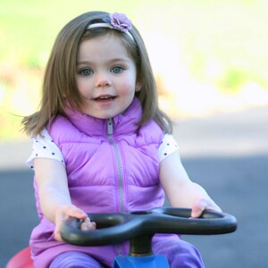 Pu&ograve; includere: Una bambina con i capelli lunghi e castani, che indossa un giubbotto viola e una camicia bianca a pois bianchi con un cerchietto di fiori, &egrave; seduta su un'auto giocattolo blu e rossa.