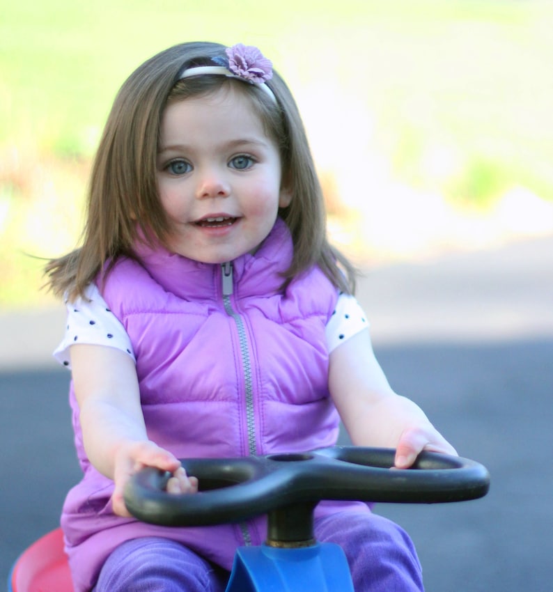 Pu&ograve; includere: Una bambina con i capelli lunghi e castani, che indossa un giubbotto viola e una camicia bianca a pois bianchi con un cerchietto di fiori, &egrave; seduta su un'auto giocattolo blu e rossa.