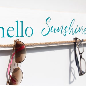 Puede incluir: Un letrero de madera blanco con el texto "hello sunshine" en letras azul turquesa. Una cuerda marr&oacute;n est&aacute; atada a trav&eacute;s del letrero con un par de gafas de sol y un par de gafas colgando.