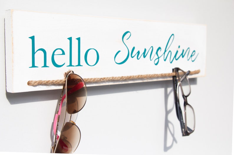 Puede incluir: Un letrero de madera blanco con el texto "hello sunshine" en letras azul turquesa. Una cuerda marr&oacute;n est&aacute; atada a trav&eacute;s del letrero con un par de gafas de sol y un par de gafas colgando.