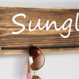 May include: Wooden sunglasses holder with the word "Sunglasses" written in white paint. A pair of sunglasses is hanging on the holder.