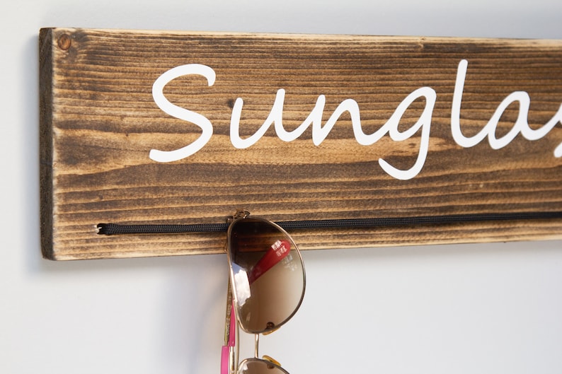 May include: Wooden sunglasses holder with the word "Sunglasses" written in white paint. A pair of sunglasses is hanging on the holder.