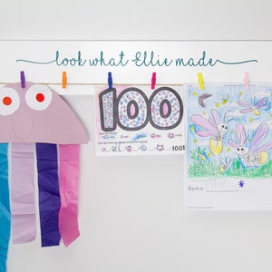 Op de afbeelding: Een wit houten bord met de tekst "look what Ellie made" hangt aan een muur. Drie kunstwerken hangen met wasknijpers aan het bord. De kunstwerken omvatten een paarse en roze kwal, een kleurrijk nummer 100 en een tekening van vuurvliegjes.