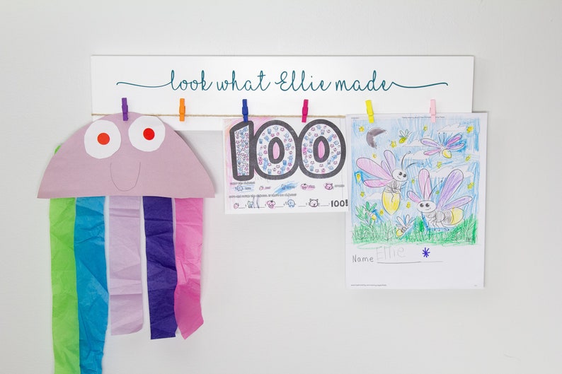 Op de afbeelding: Een wit houten bord met de tekst "look what Ellie made" hangt aan een muur. Drie kunstwerken hangen met wasknijpers aan het bord. De kunstwerken omvatten een paarse en roze kwal, een kleurrijk nummer 100 en een tekening van vuurvliegjes.