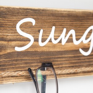 May include: A rustic wooden sign with the word "Sunglasses" written in white paint. A pair of black eyeglasses is hanging from a piece of twine attached to the sign.