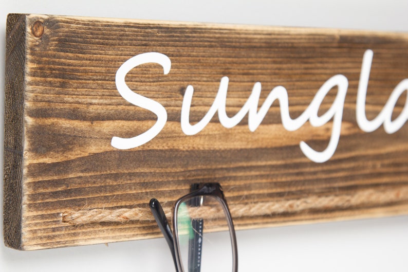 May include: A rustic wooden sign with the word "Sunglasses" written in white paint. A pair of black eyeglasses is hanging from a piece of twine attached to the sign.