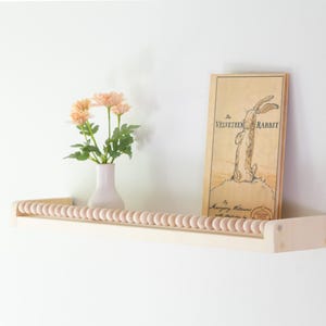 May include: A light-colored wooden shelf with a beaded front, holding a vase of peach-colored flowers and a book titled "The Velveteen Rabbit." The book has a drawing of a rabbit.