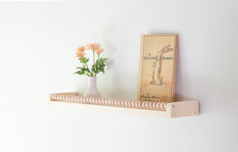 May include: A light-colored wooden shelf with a beaded front, holding a vase of peach-colored flowers and a book titled "The Velveteen Rabbit." The book has a drawing of a rabbit.