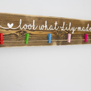 Op de afbeelding: Een rustiek houten bord met de tekst "look what lily made" in wit geschilderd. Het bord heeft een touwtje met kleurrijke wasknijpers eraan.