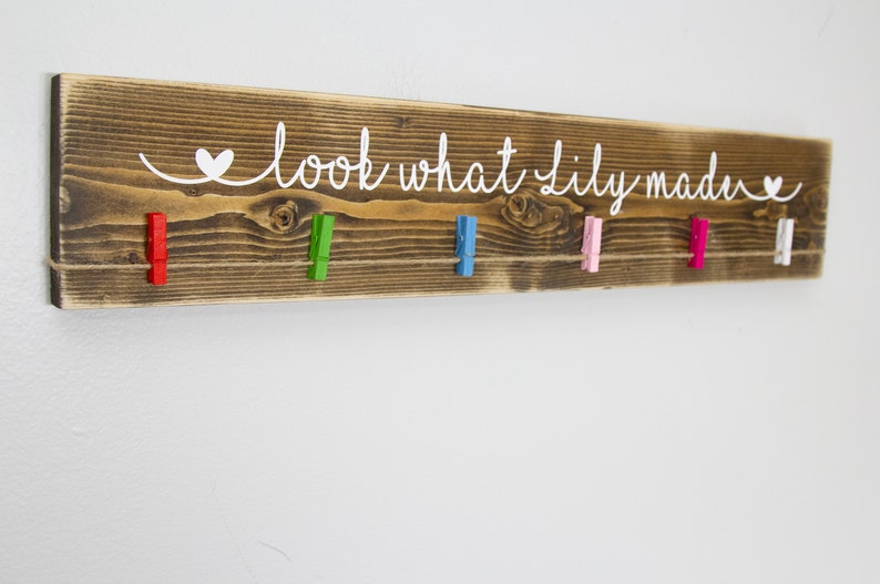 Op de afbeelding: Een rustiek houten bord met de tekst "look what lily made" in wit geschilderd. Het bord heeft een touwtje met kleurrijke wasknijpers eraan.