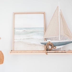 May include: A framed watercolor print of a beach scene and a decorative sailboat on a wooden shelf. The sailboat has a blue hull, beige sails, and a rope detail. The shelf is adorned with small wooden beads.