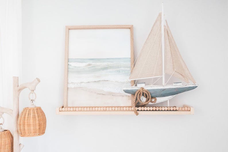 May include: A framed watercolor print of a beach scene and a decorative sailboat on a wooden shelf. The sailboat has a blue hull, beige sails, and a rope detail. The shelf is adorned with small wooden beads.