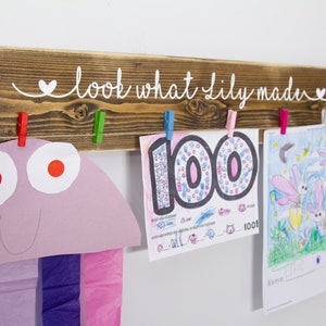 Op de afbeelding: Een houten bord met de tekst "look what Lily made" in witte verf. Het bord heeft vijf wasknijpers die kunstwerken vasthouden, waaronder een tekening van vuurvliegjes en een werkblad met het getal 100.