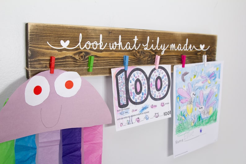 Op de afbeelding: Een houten bord met de tekst "look what Lily made" in witte verf. Het bord heeft vijf wasknijpers die kunstwerken vasthouden, waaronder een tekening van vuurvliegjes en een werkblad met het getal 100.