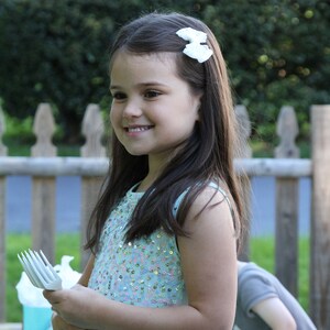 Pu&ograve; includere: Una bambina con un vestito blu chiaro con paillettes e un fiocco bianco nei capelli tiene una forchetta di plastica bianca. Sta sorridendo e guardando la telecamera.