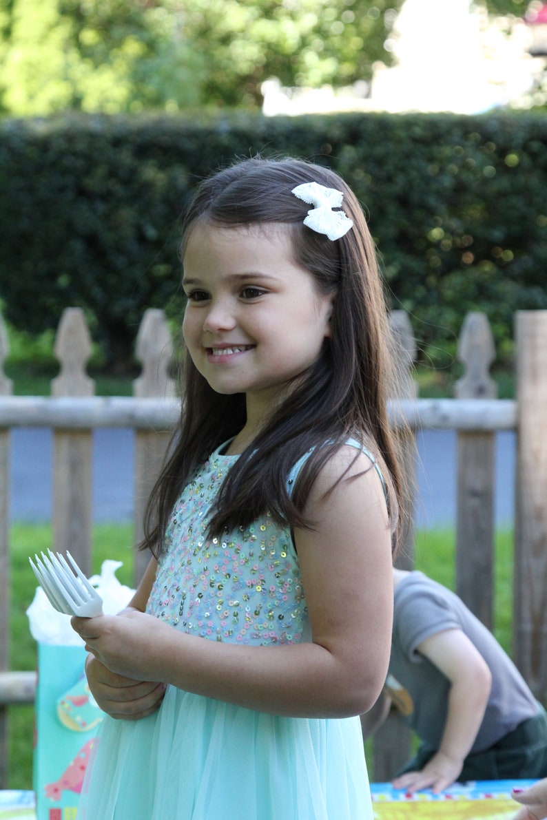 Pu&ograve; includere: Una bambina con un vestito blu chiaro con paillettes e un fiocco bianco nei capelli tiene una forchetta di plastica bianca. Sta sorridendo e guardando la telecamera.