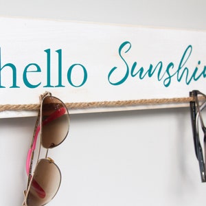 Puede incluir: Un letrero de madera blanco con el texto "hello sunshine" en letras azul turquesa. Una cuerda marr&oacute;n est&aacute; atada a trav&eacute;s del letrero con gafas de sol colgando.