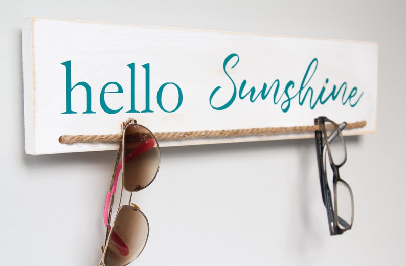 Puede incluir: Un letrero de madera blanco con el texto "hello sunshine" en letras azul turquesa. Una cuerda marr&oacute;n est&aacute; atada a trav&eacute;s del letrero con gafas de sol colgando.