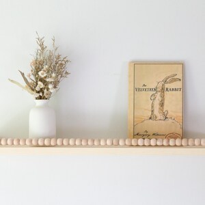 May include: A wooden shelf with a row of wooden beads, holding a white vase with dried flowers and a vintage copy of "The Velveteen Rabbit" book. The book has a rabbit illustration.