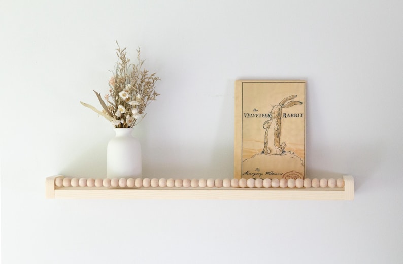 May include: A wooden shelf with a row of wooden beads, holding a white vase with dried flowers and a vintage copy of "The Velveteen Rabbit" book. The book has a rabbit illustration.