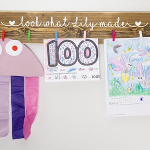 Op de afbeelding: Een houten bord met de tekst "look what lily made" in witte verf. Het bord heeft aan elke kant van de tekst een hartje. Er zijn vijf wasknijpers aan het bord bevestigd, die drie kunstwerken vasthouden. De kunstwerken omvatten een kleurrijke tekening van vuurvliegjes, een tekening van het getal 100 en een papieren knutselwerk van een kwal.