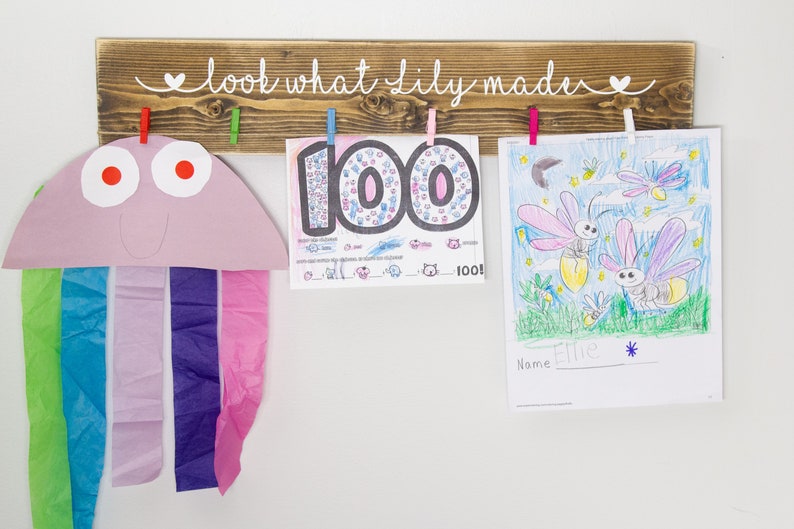 Op de afbeelding: Een houten bord met de tekst "look what lily made" in witte verf. Het bord heeft aan elke kant van de tekst een hartje. Er zijn vijf wasknijpers aan het bord bevestigd, die drie kunstwerken vasthouden. De kunstwerken omvatten een kleurrijke tekening van vuurvliegjes, een tekening van het getal 100 en een papieren knutselwerk van een kwal.