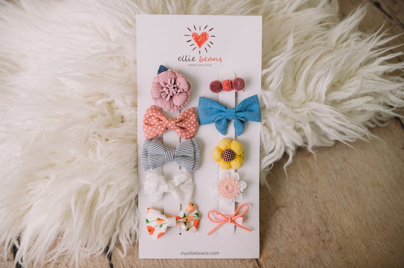 Pu&ograve; includere: Un set di otto mollette per capelli con vari design, tra cui fiori, fiocchi e pois. Le mollette sono disposte su una carta bianca con il testo "ellie beans made with love" e "myelliebeans.com".