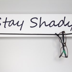 May include: White wooden sign with black cursive text that reads "Stay Shady". A pair of black and green glasses hangs from a black string attached to the sign.