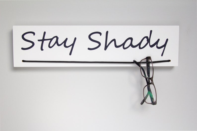 May include: White wooden sign with black cursive text that reads "Stay Shady". A pair of black and green glasses hangs from a black string attached to the sign.